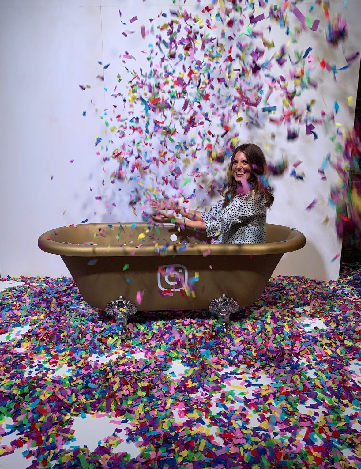 A woman in a silver patterned robe sits inside a vintage-style gold bathtub surrounded by a massive shower of colorful confetti, with confetti covering the floor and splashing high into the air against a white studio backdrop.

