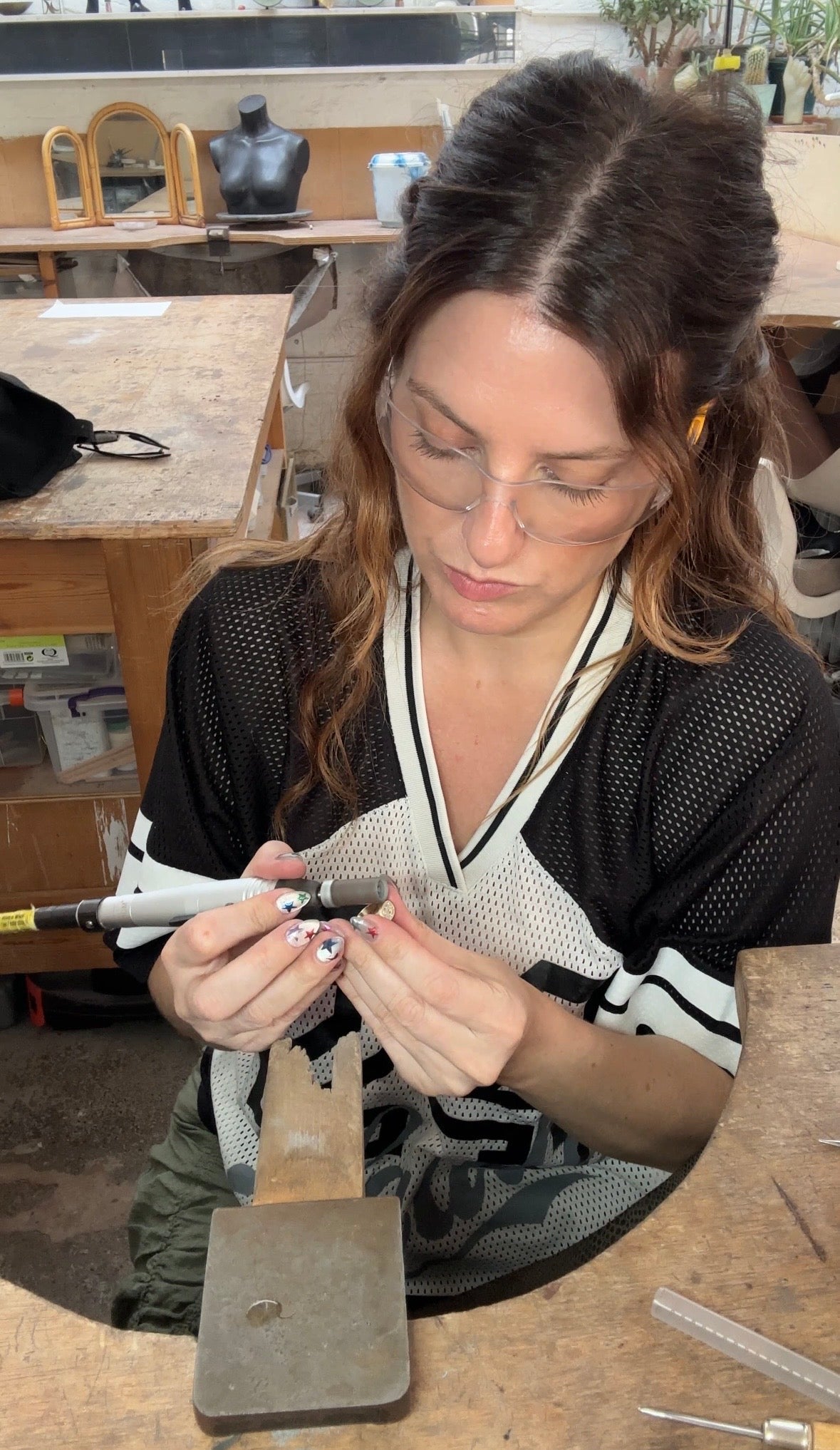 A woman wearing glasses and a black-and-white mesh top is seated at a wooden table, using a small hand tool on a metal piece held in a clamp. The setting appears to be a workshop or studio with tools and workbenches visible in the background.

