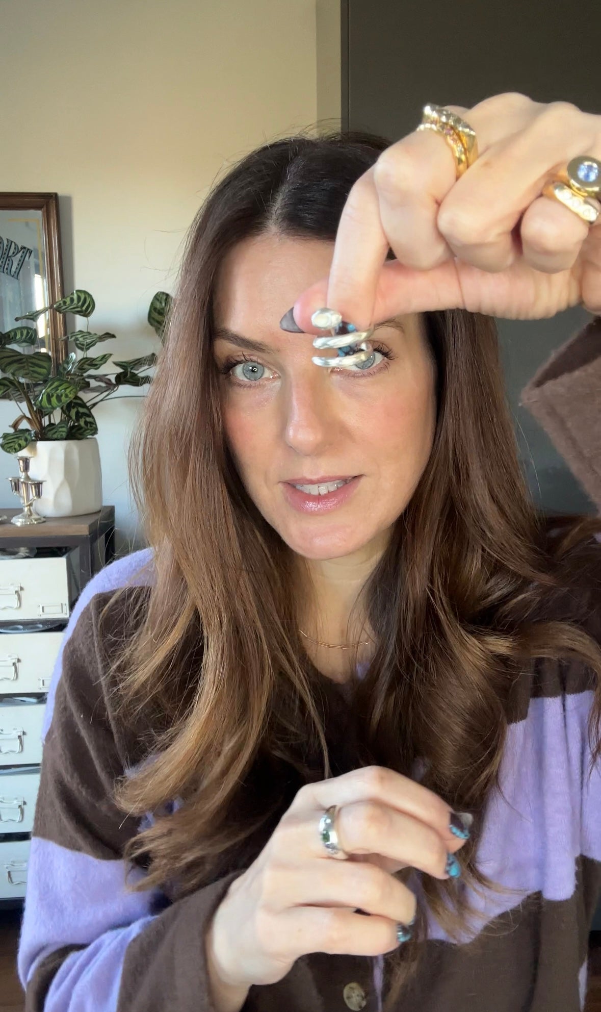 A woman with long brown hair holds up a pair of silver rings with black and white patterned bands, showing them close to the camera. She is wearing other rings on her fingers and has a purple and brown striped shirt in a home setting.

