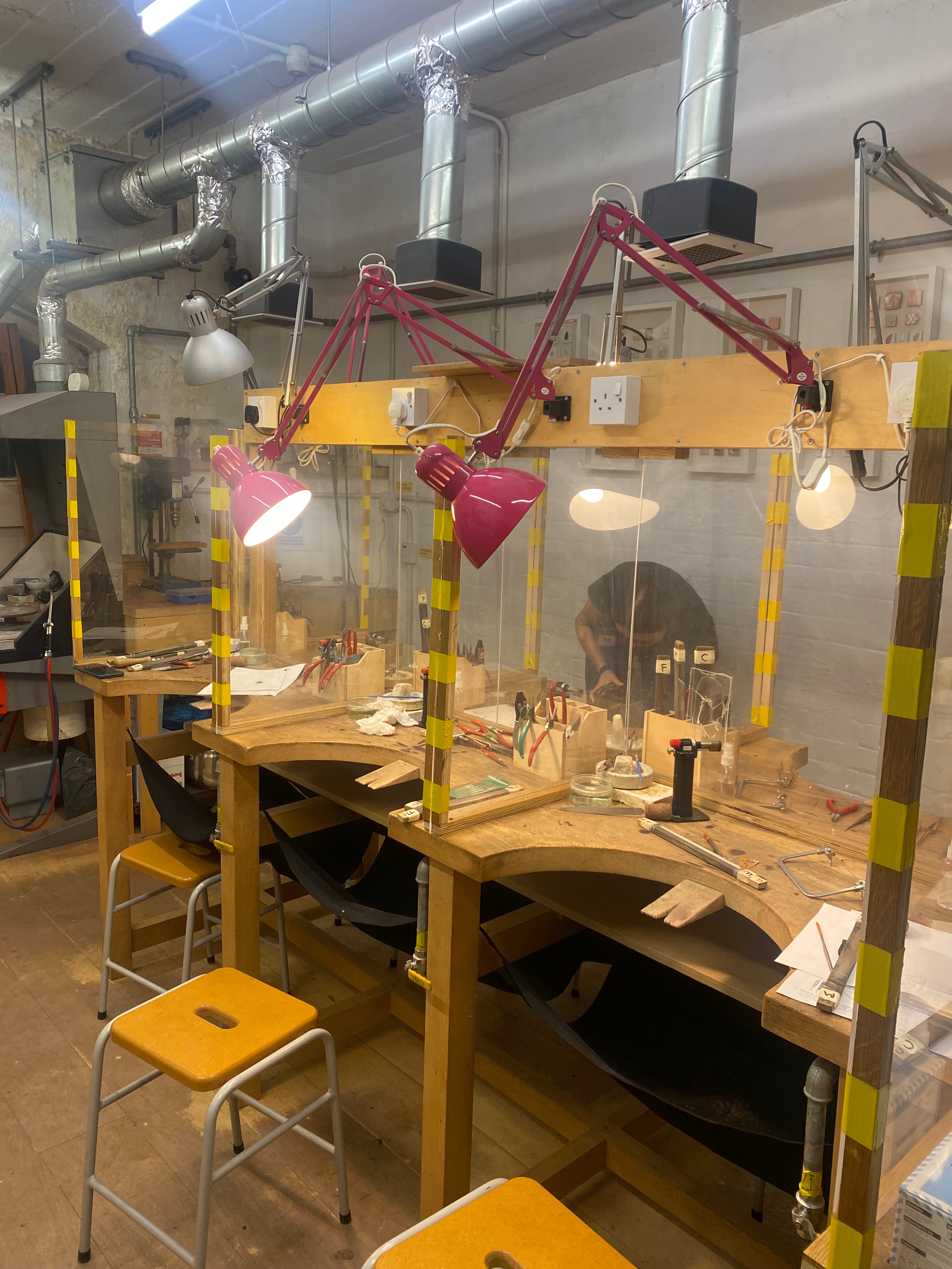 A workshop station with a wooden workbench, clear protective guard panels, adjustable magenta and silver work lights, and yellow-and-black striped safety tape. Tools and small equipment are arranged on the bench, with stools and industrial ceiling ducts visible in the room.

