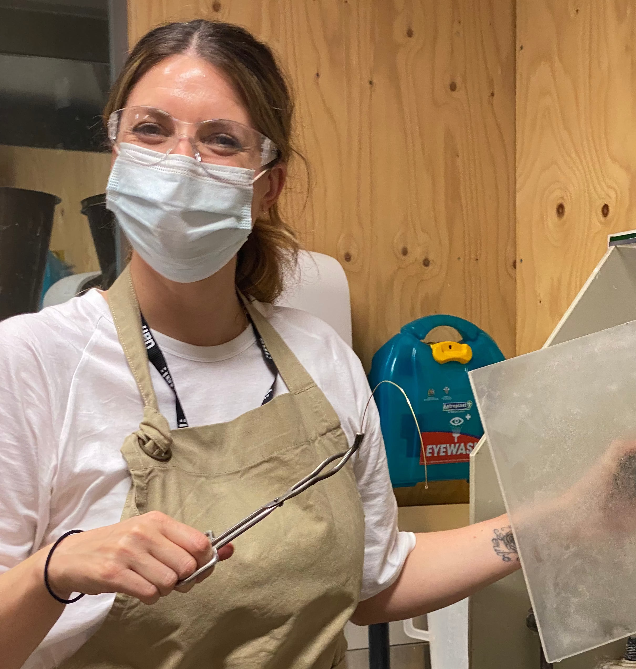 A woman wearing a beige canvas apron over a white t-shirt and a light blue face mask stands in a workshop setting, holding a clear glass lid next to a metal machine.

