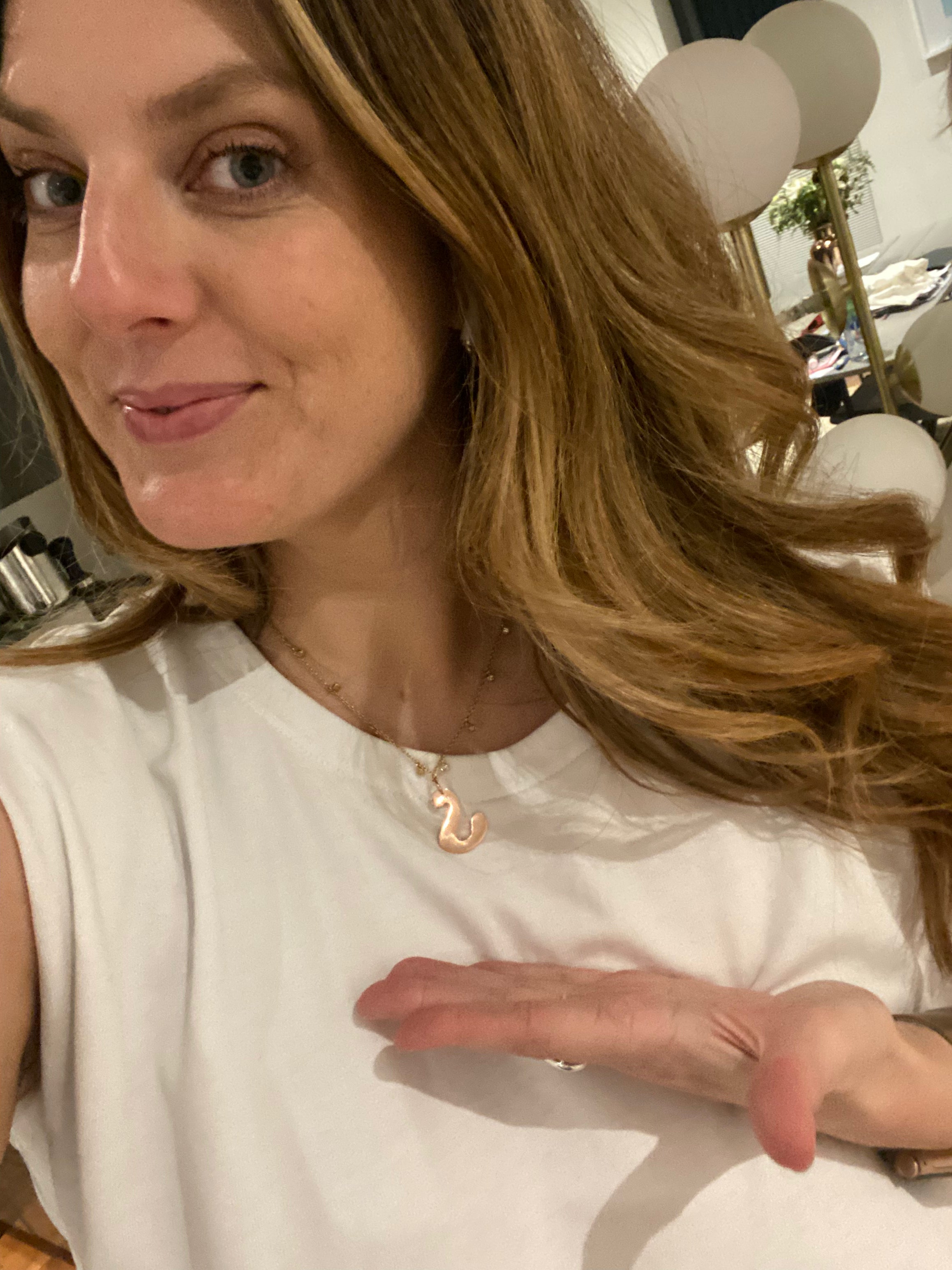 A woman with long light brown hair wearing a white sleeveless top and a delicate rose gold necklace with a small pendant, posing indoors.

