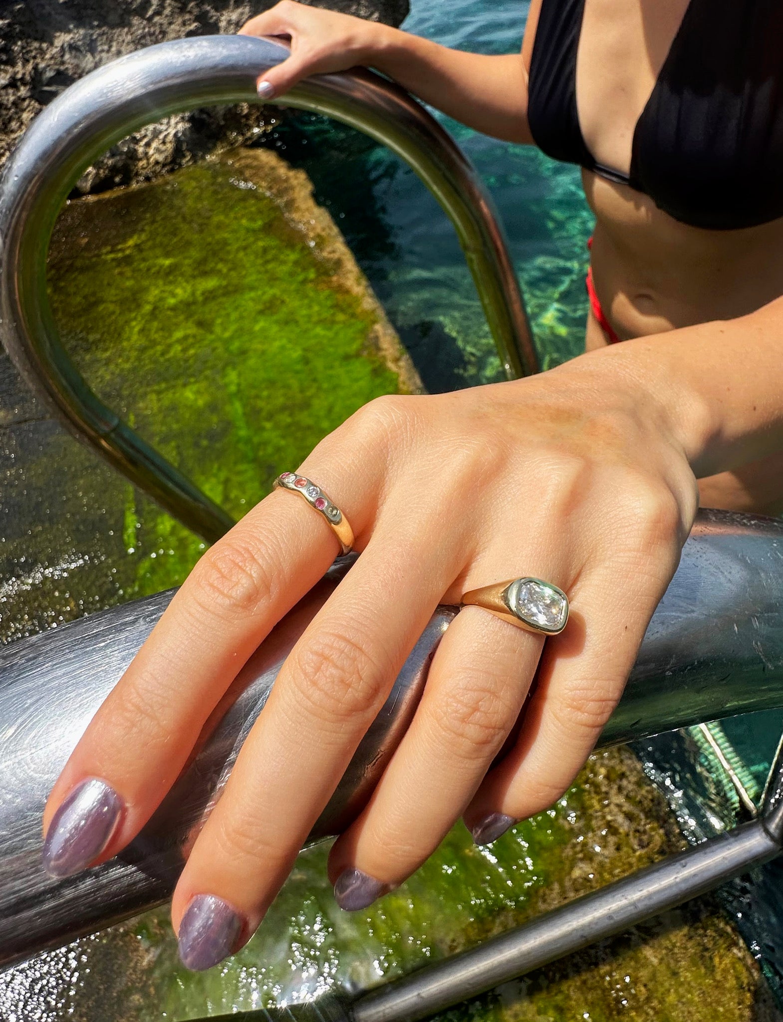 A close-up lifestyle shot of a person's hand resting on a metal pool railing, showcasing two rings. One ring is a simple gold band with small clear stones, and the other is a gold ring with a large oval clear gemstone.

